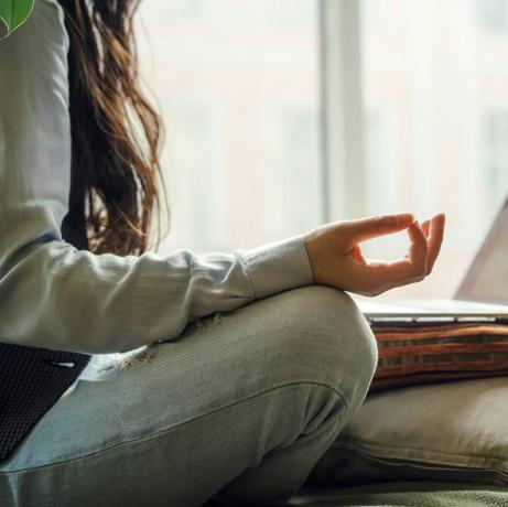 Mavi kot pantolonlu orta yaşlı Asyalı bir kadın, dizüstü bilgisayarın önünde yoga pozu vererek yatakta oturuyor, evde uzaktan çalışıyor, karantina döneminde internetten alışveriş yapıyor.