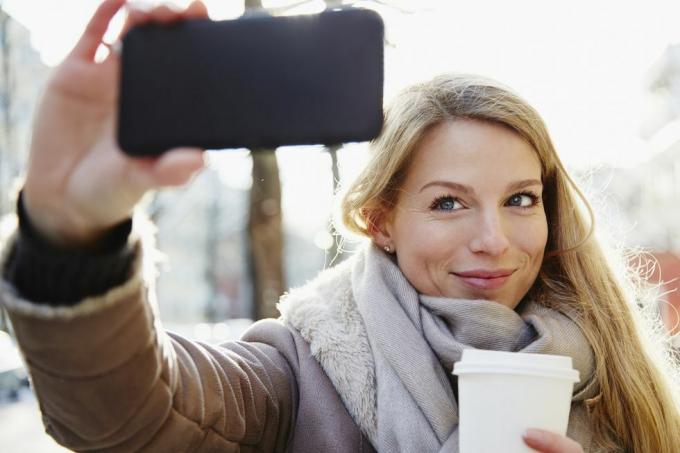 Kışın şehrin sokaklarında yürüyen yakışıklı kadın selfie çekiyor ve elinde kahve tutuyor, arkadan aydınlatmalı