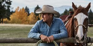 Kevin Costner Yellowstone'da, elinde iple çite yaslanmış, soluk mavi bir kot gömlek ve bej kovboy şapkası takmış bir atın yanında.