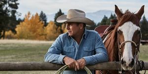 Kevin Costner Yellowstone'da, elinde iple çite yaslanmış, soluk mavi bir kot gömlek ve bej kovboy şapkası takmış bir atın yanında.