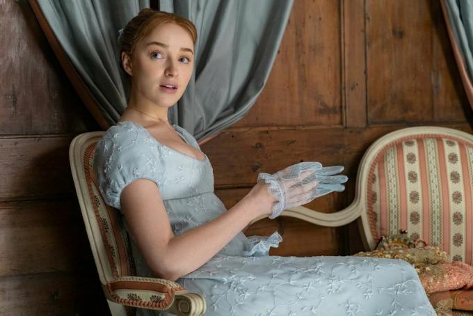 phoebe dynevor 102. bölümde daphne bridgerton rolünde, oturmuş, mavi, imparatorluk belli bir elbise giyiyor