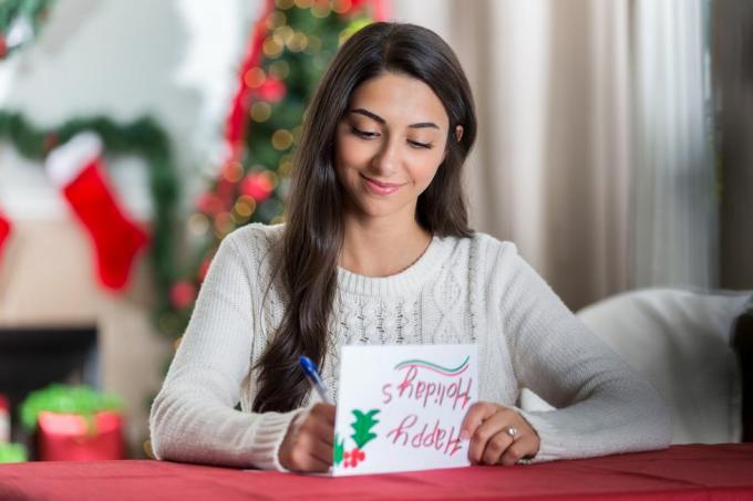Gülümseyen genç bir kadın, dekore edilmiş oturma odasında bir masada oturuyor ve bir arkadaşına Noel için bir mektup yazarken konsantrasyonla aşağıya bakıyor.